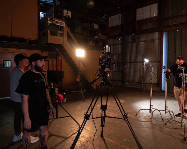 A camera is set up on a tripod in a dimly lit warehouse, with two individuals watching and a third person standing by a lighting setup.