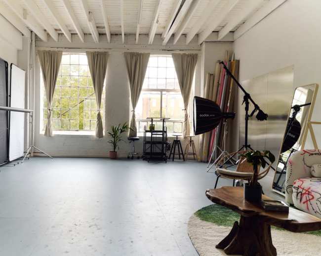 The image depicts a spacious photography studio featuring large windows, equipment like light stands and a reflector, a potted plant, and a few pieces of furniture.