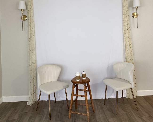 The image shows two white chairs on either side of a small wooden stool in front of a plain white backdrop draped with fabric and wall sconces.
