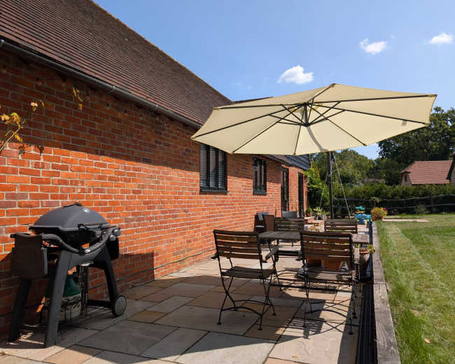 A patio features a dining set with chairs and a table, an umbrella for shade, and a grill, situated against a brick wall of a house.