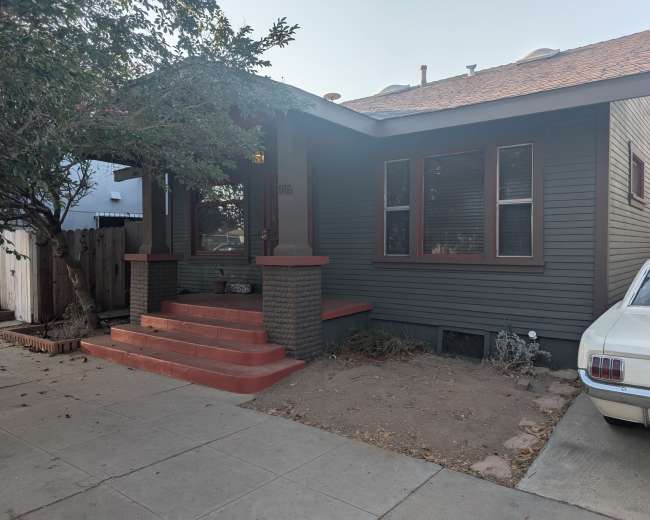 The image shows a single-story house with dark green siding, red trim, and a concrete walkway leading to the front porch, alongside a vintage white car parked on the side.