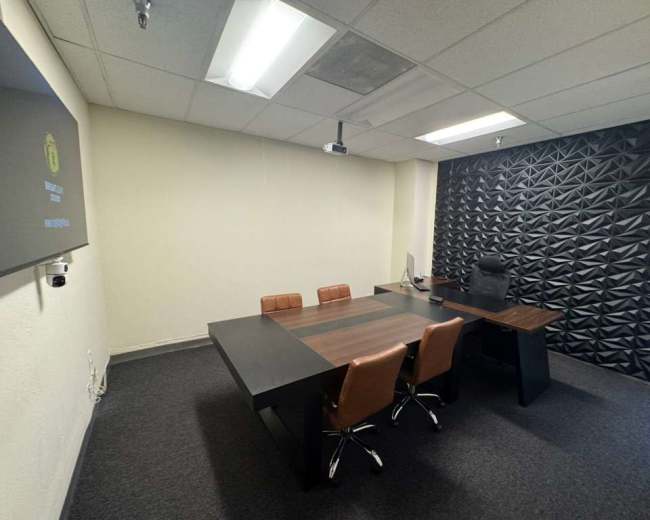 The image shows a modern office with a conference table surrounded by four chairs, a computer workstation, and a textured black wall.
