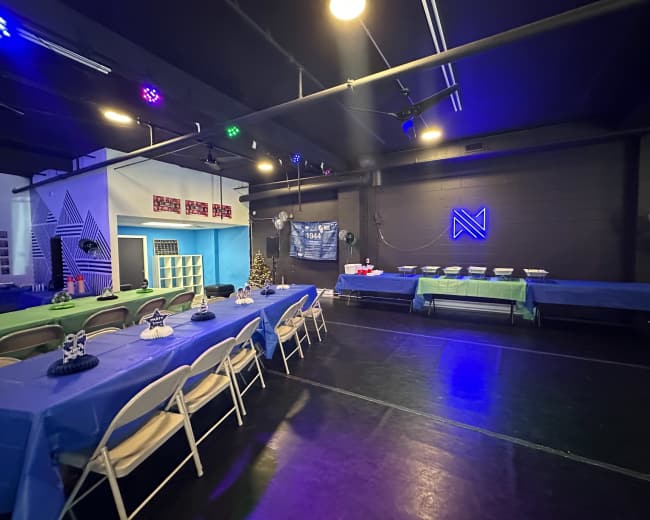 The image shows a decorated event space with tables covered in blue cloth, arranged for a gathering, and features a snack table against the wall.
