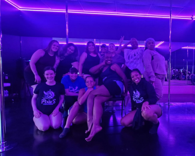 A group of eight individuals poses together in a dimly lit dance studio with purple neon lighting and a mirrored wall.