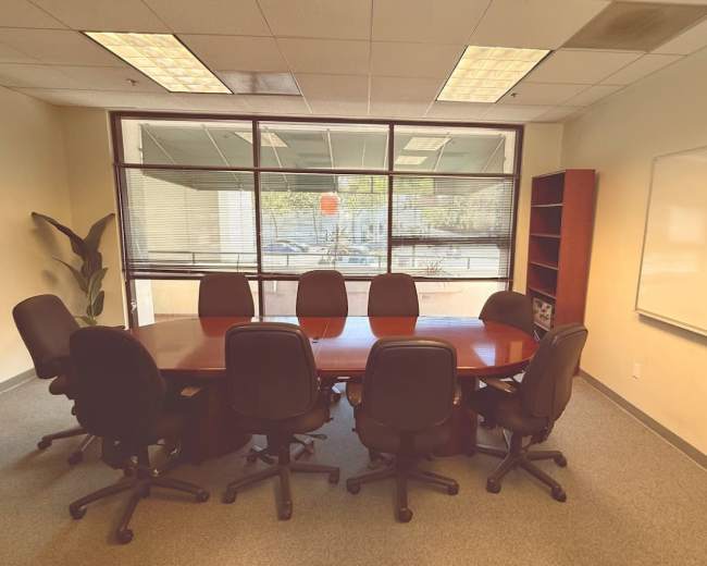 A conference room features a large oval table surrounded by eight black chairs, with windows providing natural light and a whiteboard on one wall.