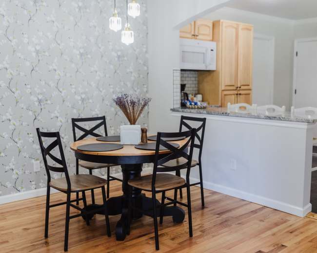 A small dining area features a round table with four black chairs, surrounded by floral wallpaper and adjacent to a kitchen with wooden cabinetry and a bar counter.