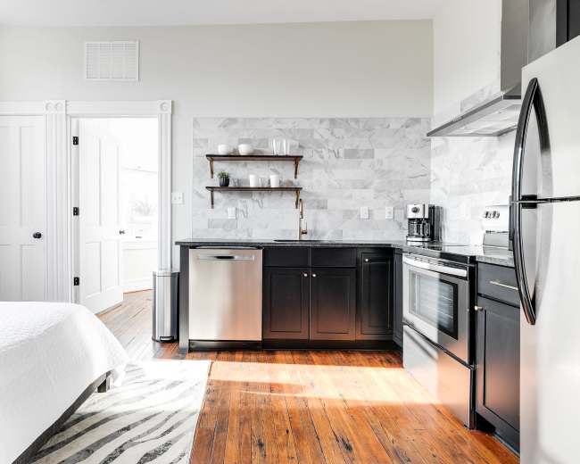 The image shows a modern studio apartment featuring a bed in the foreground, a kitchen area with a stainless steel refrigerator and oven, and a door leading to another room.