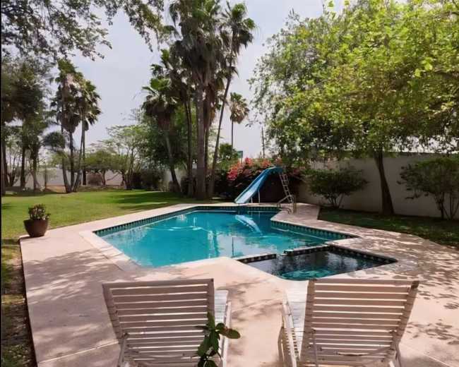 The image shows a rectangular swimming pool with a slide, surrounded by grass and palm trees, alongside a pair of white lounge chairs.