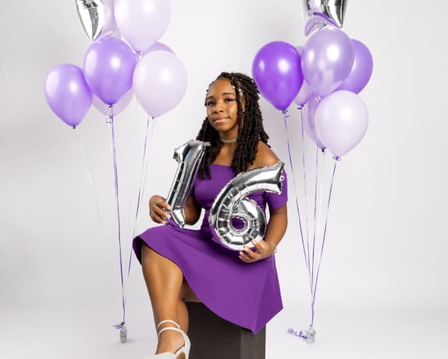 A girl in a purple dress sits on a black cube, holding a silver "16" balloon, surrounded by purple and silver balloons.