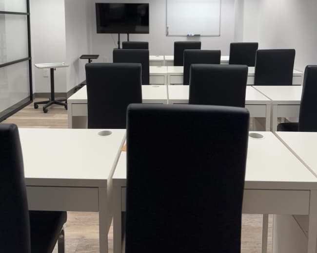 The image shows a modern classroom with rows of white desks and black chairs, featuring a television and a whiteboard at the front.