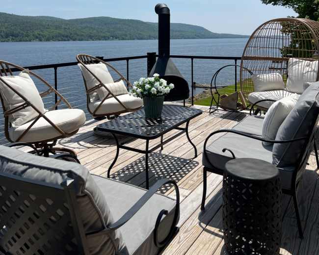 The image shows a patio by a river, featuring several chairs, a coffee table, and a large decorative planter, with a scenic view of green hills in the background.