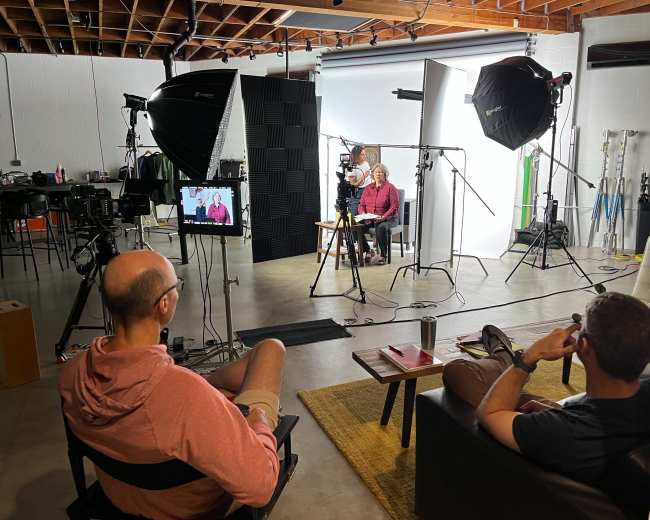 A studio setting with two men seated in front of camera equipment, lighting, and a monitor displaying an interview being filmed.