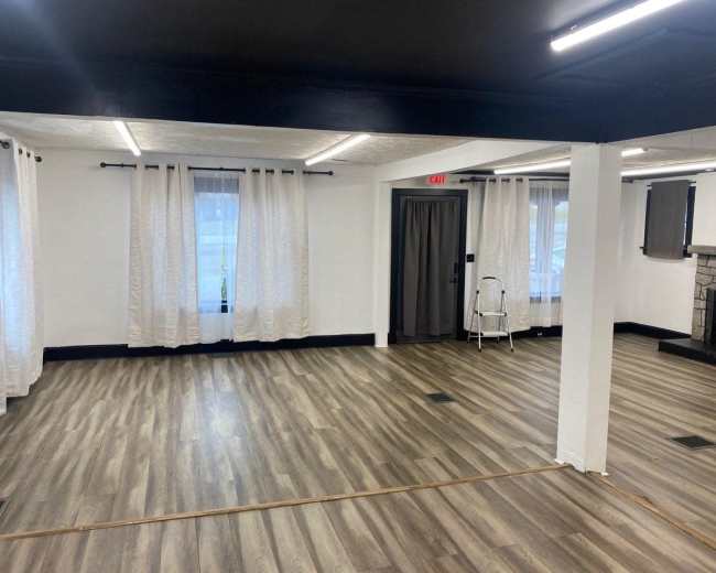 The image shows a spacious room with light-colored wooden flooring, large windows covered with white curtains, and minimal furnishings.