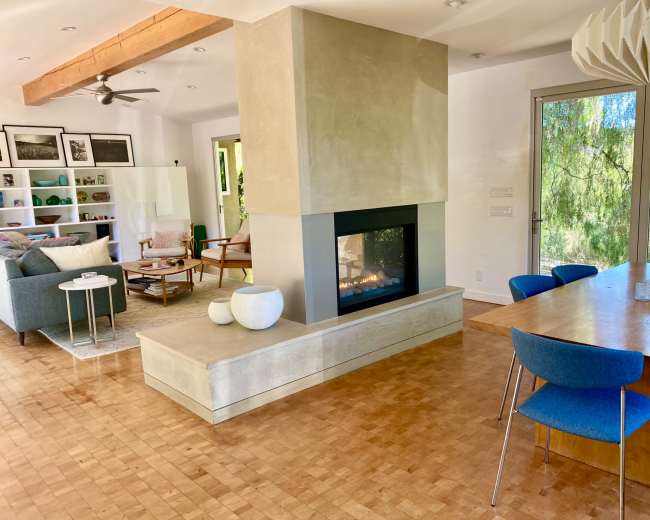 The image shows a modern living space featuring a central fireplace, dining area, and a seating area with large windows overlooking greenery.