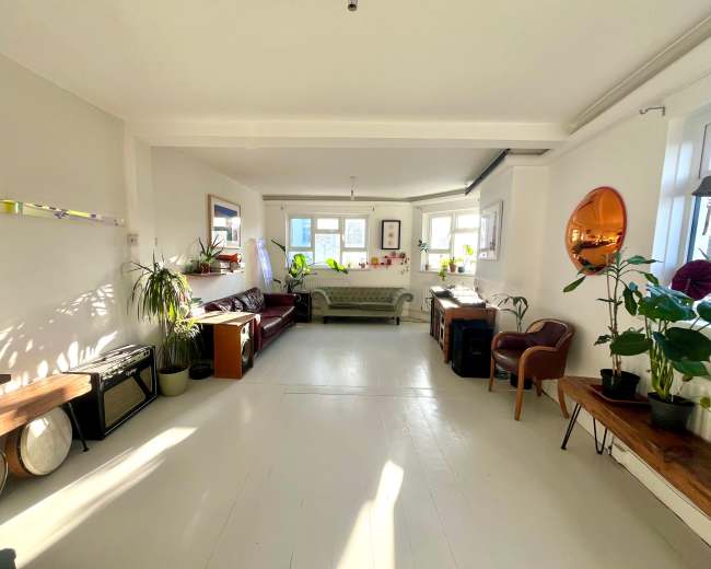 The image shows a bright, open living space featuring a mix of seating areas, plants, and natural light coming through the windows.