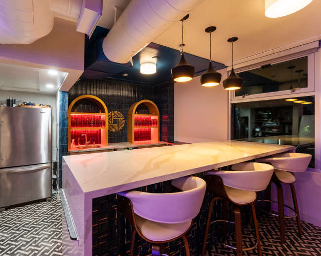 The image shows a modern kitchen and bar area with a sleek countertop, stylish bar stools, and ambient lighting highlighting the backlit shelves.