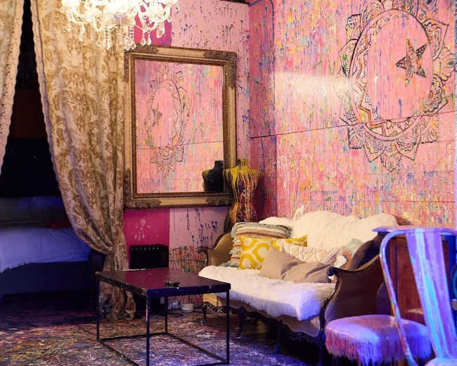 The scene features a colorful, artistically painted room decorated with a chandelier, ornate mirror, and a mix of vintage and modern furniture.