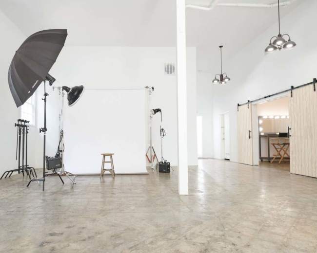 The image shows a spacious photography studio with a simple backdrop, lighting equipment, and a stool set against a light-colored wall.