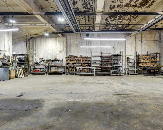 The image shows an industrial workshop with concrete flooring, exposed walls, and metal shelves filled with wooden boxes and tools.