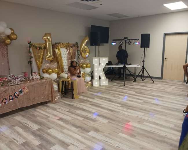 A child sits on a chair in front of a decorated table with a "16" sign and party supplies in a reception hall.