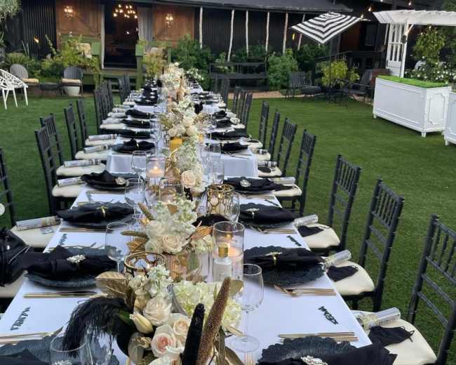 A long outdoor dining table is set with black and white tableware, decorated with floral arrangements and candles, in a garden setting.