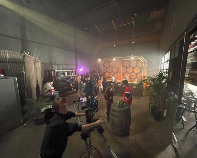 A group of people is gathered in a dimly lit warehouse setting with barrels and a backdrop of large circular decorations.