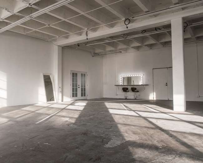 An empty room with a concrete floor, large windows casting shadows, a mirror leaning against the wall, and a vanity with two chairs.