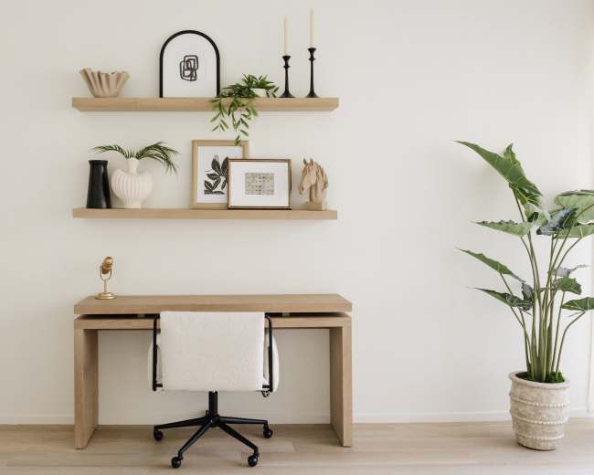 A minimalist workspace features a wooden desk with a chair, flanked by decorative shelves that display various objects and plants against a plain white wall.