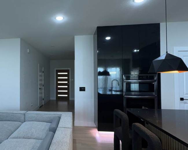 The image shows a modern, open-concept living space featuring a light-colored sofa, a sleek kitchen with black cabinetry, and a view of a hallway with a wooden door.