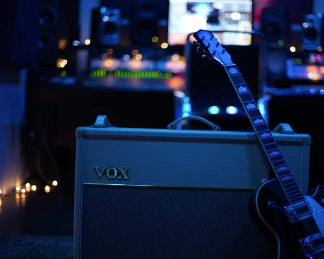 A black electric guitar leans against a Vox amplifier in a dimly lit recording studio filled with audio equipment.