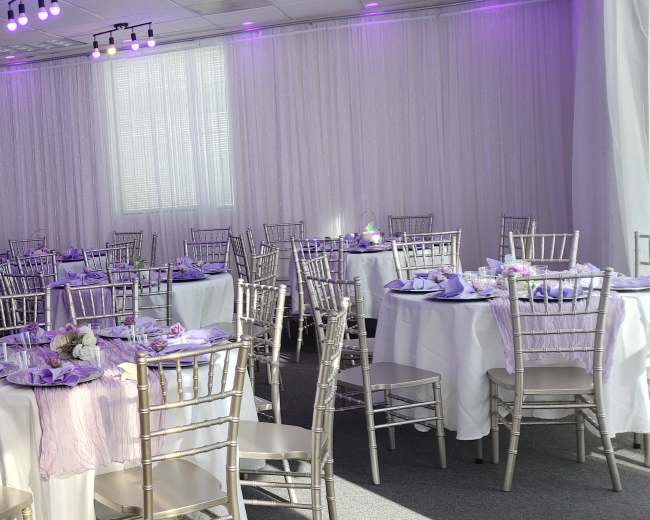 The image shows a banquet hall set up with several round tables covered in white tablecloths, adorned with purple accents and silver chiavari chairs.