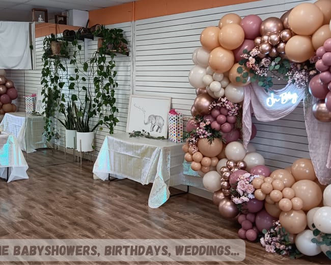 The image shows a decorated indoor space featuring tables with desserts and a balloon arch adorned with flowers, designed for events like baby showers and birthdays.