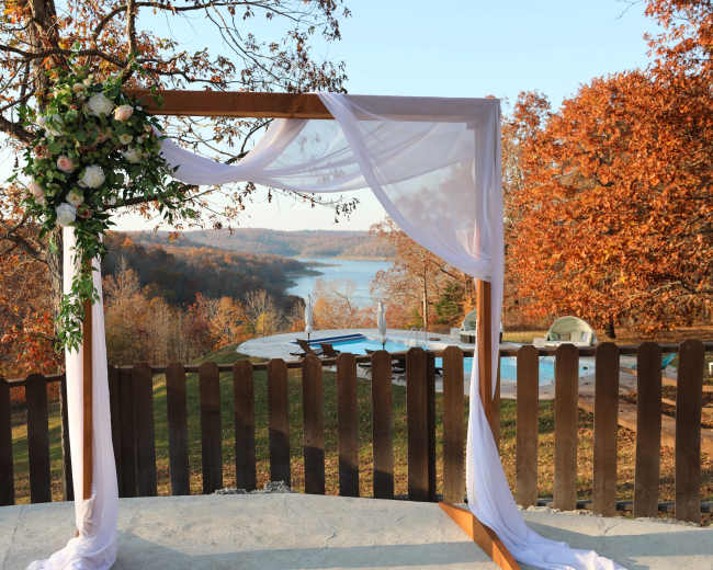 A wooden arbor draped with white fabric and flowers overlooks a lake surrounded by autumn foliage and a swimming pool.