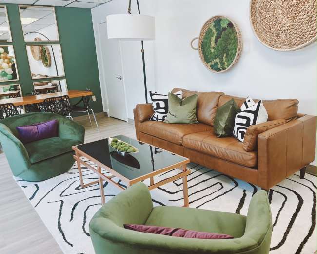 A stylish living room with a brown leather sofa, two green chairs, a glass coffee table, and wall decor featuring woven baskets.