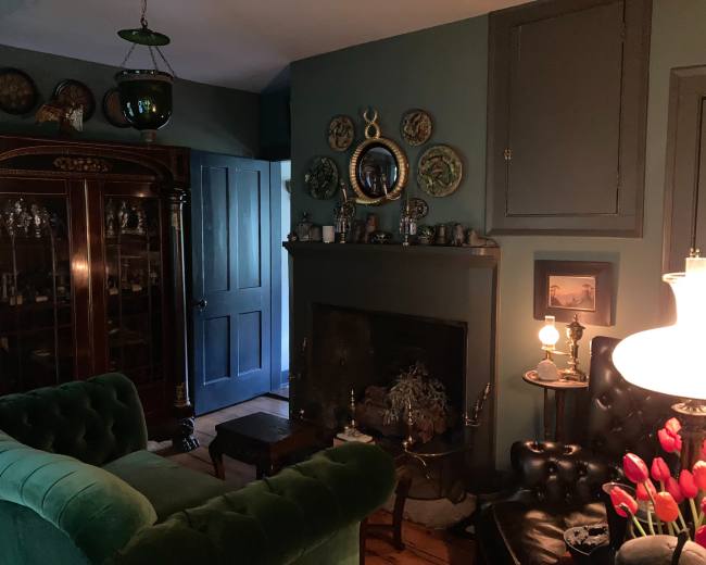 The room features a dark green color scheme with a vintage cabinet, a fireplace adorned with various items, and plush seating arranged around a small table.