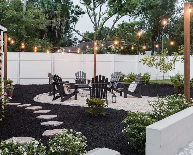 A circular seating area with Adirondack chairs is surrounded by decorative gravel and lined with stepping stones, illuminated by string lights in a landscaped backyard setting.