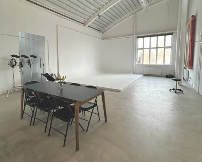 The image shows a spacious, well-lit studio with a large white wall, a small white platform, and a black table with chairs.