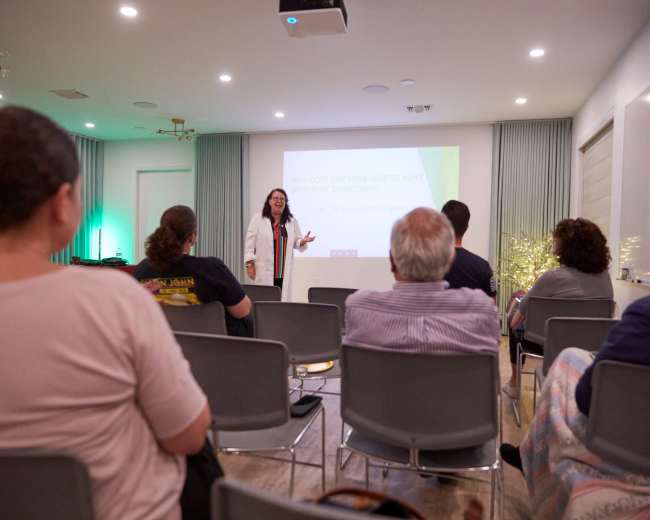 A presenter stands in front of an audience seated in a room with a projection screen, delivering a lecture on the benefits of a certain treatment for various conditions.