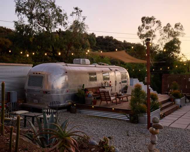 An Airstream trailer is parked on a wooden deck in a landscaped area with string lights and various plants, against a backdrop of hills at sunset.