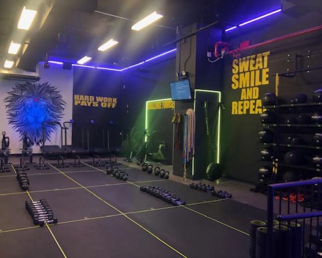 The image shows a brightly lit gym interior featuring various workout equipment, including dumbbells and resistance bands, with motivational phrases painted on the walls.