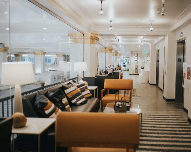 The image shows a well-lit hallway with modern furniture, including black sofas and orange chairs, alongside framed artwork and decorative elements on the walls.