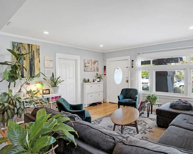 The living room features a sectional sofa, two green armchairs, a round coffee table, and various plants, with natural light streaming in through a large window.