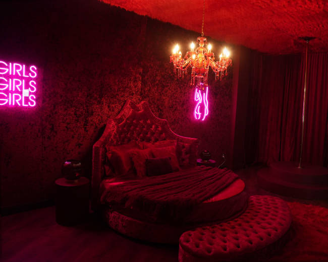 The room features a round bed with a velvet headboard, illuminated by a chandelier and a neon sign, with red walls and a pole in one corner.