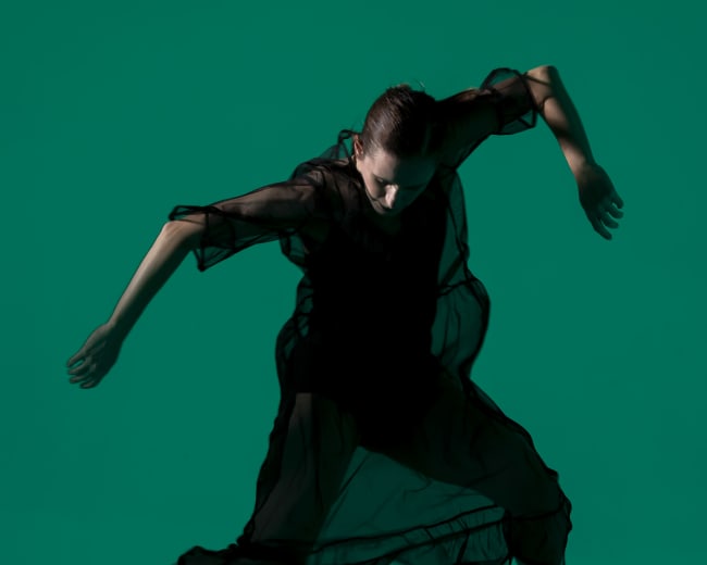 A dancer in a black, sheer outfit leaps against a solid teal background.