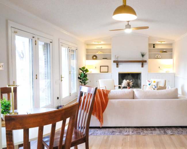The image shows a bright living room featuring a white sofa, a fireplace with shelving, and large windows leading to an outdoor space.