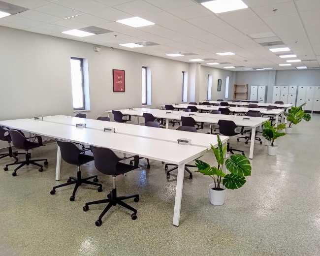 A spacious, modern office area with rows of white tables and black chairs arranged neatly, alongside potted plants and lockers.