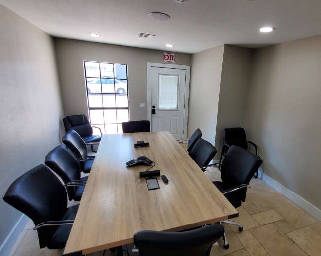 A conference room features a large wooden table surrounded by black chairs and a wall with a windowed door leading outside.