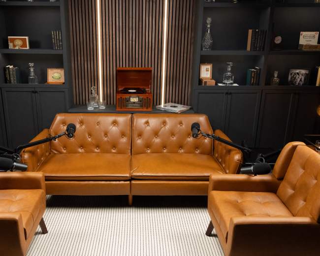 The image shows a cozy podcast studio featuring two brown leather sofas with microphones, surrounded by shelves lined with books and decorative items.