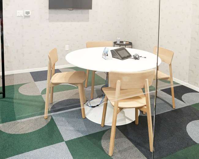 The image shows a small meeting room with a round table, four chairs, and a mounted television on the wall, featuring a modern design and colorful carpeting.