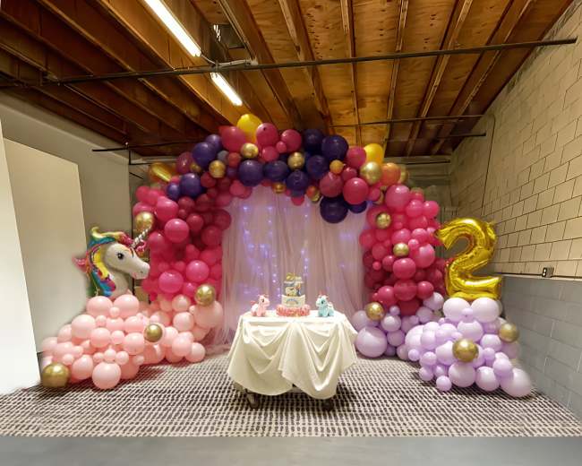 The image shows a decorative setup for a birthday party featuring a table with a cake, surrounded by an arch of colorful balloons and a backdrop.